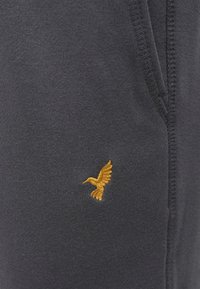 Sötétszürke szövet lágy tapintással, arany hímzett madárlogóval a zsebrész közelében. Látható varrási részleteket tartalmaz.