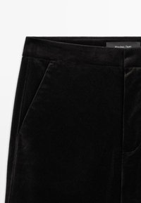 Pantalon en velours noir avec poche avant inclinée et ceinture, avec une étiquette Massimo Dutti partiellement visible.