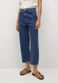 Jeans de mezclilla azul de tiro alto con un ajuste relajado y recortado, que cuentan con un cierre frontal de botón y un diseño clásico de cinco bolsillos, combinados con sandalias marrones.