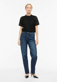 Haut en dentelle noire à manches courtes, col à volants, associé à un jean bleu foncé taille haute et des escarpins noirs à bout pointu.