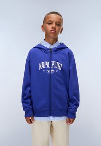 Sudadera azul con cremallera y logo delantero, con un ajuste relajado, mangas largas y capucha con cordón, superpuesta a una camisa de cuello azul claro.