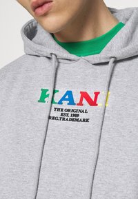Felpa grigia chiara in cotone, con cappuccio regolabile. Sul fronte è stampato un testo multicolore: "KANI" e "THE ORIGINAL EST. 1989."