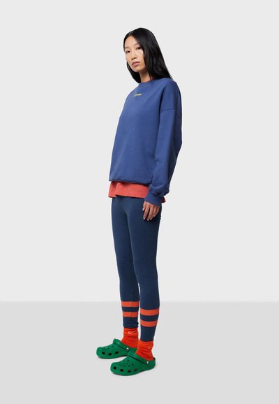 Marinblå oversized sweatshirt, korallfärgad underlayer, mörkblå leggings med oranga ränder, gröna träskor, oranga strumpor. Mjuka tygtexturer.