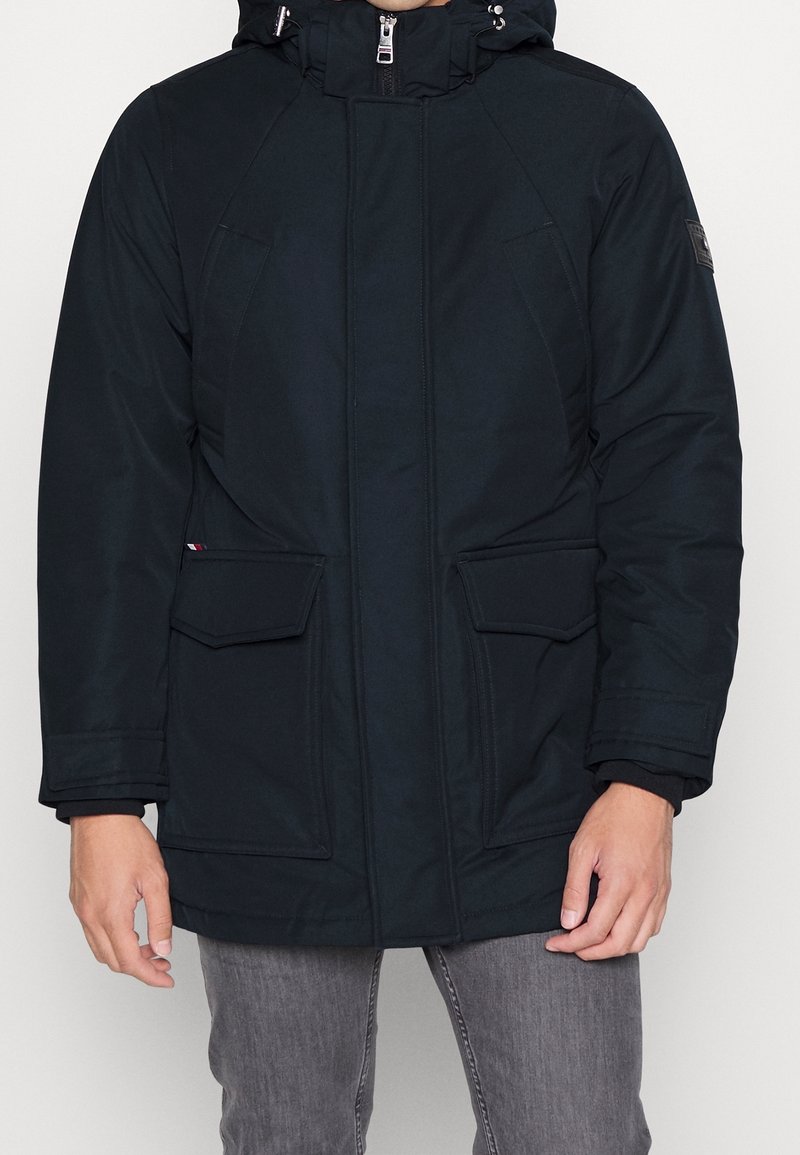 Černá zateplená parka se zapínáním na zip, nastavitelnou kapucí a dvěma velkými předními kapsami. Vyrobena z hladkého, voděodolného materiálu.