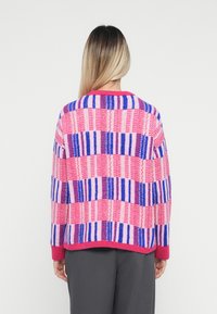 Person with long hair wearing a pink, purple, and red striped sweater and gray pants, facing away against a white background.