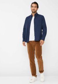 Marineblaues Hemd mit Knöpfen und Tasche, weißes T-Shirt, braune Cordhosen und weiße Sneakers. Das Outfit hat einen lässigen Schnitt und glatte Texturen.