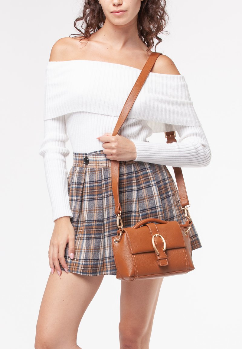 Borsa a tracolla in pelle marrone con un design strutturato, accessori dorati e un manico superiore. Il modello indossa una maglietta bianca a coste off-shoulder e una gonna a quadri.