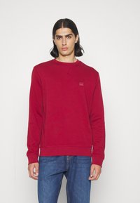 Felpa rossa a girocollo realizzata in materiale morbido, con polsini e orlo a coste, e una piccola etichetta con logo sul petto. Indossata con jeans blu.