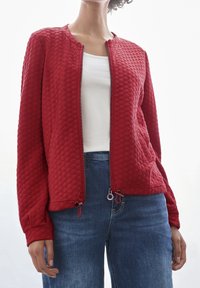 Chaqueta acolchada roja con cuello redondo, cierre frontal completo y dobladillo con cordón. Presenta mangas largas y un acabado de tela texturizado.