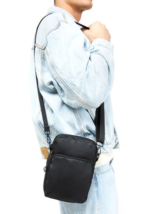 Black crossbody bag with a textured surface, two main compartments, adjustable strap, and metal hardware, worn over a light denim jacket.