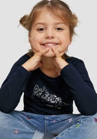 Marineblauwe longsleeve shirt met een wit bloemen- en tekstontwerp, gecombineerd met lichtblauwe jeans met kleurrijke geborduurde accenten. Kind dat glimlacht.