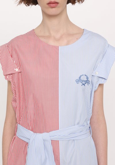 Chemise à rayures rouges et bleues à manches courtes, avec une ceinture à la taille et un détail de logo brodé en bleu sur la section bleue.