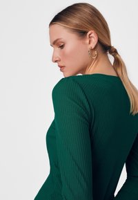 Zielona, prążkowana bluzka z długim rękawem i okrągłym dekoltem. Tekstura materiału jest wyraźnie widoczna. Złoty kolczyk w kształcie koła widoczny na uchu modelki.