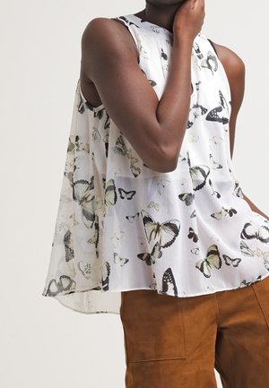 Mouwloze witte blouse met zwart en bleekgele vlinderprint, gedragen met een bruine broek, persoon poseert met hand dichtbij het gezicht.