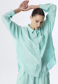Camisa de color verde menta de corte suelto, hecha de un tejido texturizado, con un cierre de botones en la parte delantera, mangas largas y un pequeño bolsillo en el lado izquierdo.