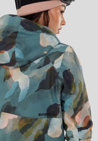 Žena v mnohobarevné maskáčové bundě s kapucí a růžovou čepicí, stojící bokem na neutrálním šedém pozadí.