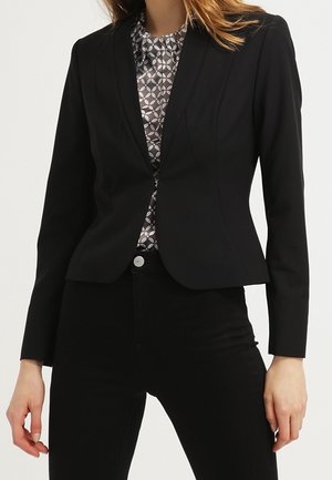 Kvinde iført en tætsiddende sort blazer over en mønstret grå og hvid bluse, kombineret med sorte bukser med højtalje.