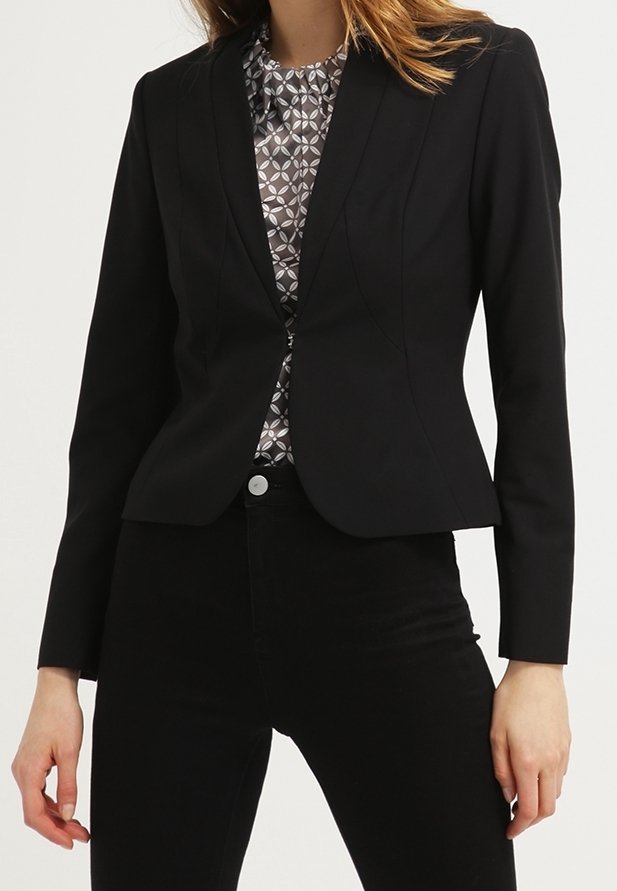 Femme portant un blazer noir cintré sur une blouse grise et blanche à motifs, assorti à un pantalon noir taille haute.