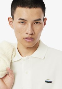 Witte poloshirt van een textuurstof, met een meerkleurig geborduurd krokodillogo op de linkermouw. Model houdt een crème trui vast.