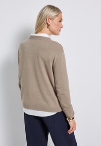 Beige knit sweater with a ribbed hem and cuffs, featuring a casual fit. Worn over a white collared shirt, with a smooth texture.