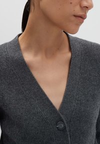 Femme portant un cardigan en tricot côtelé gris foncé avec un décolleté en V profond et un bouton visible, vu du cou à la partie supérieure de la poitrine.