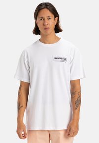 Camiseta de algodón blanca con un corte relajado, que presenta un pequeño logo rectangular de Quiksilver en el lado izquierdo del pecho. Manga corta y cuello redondo.