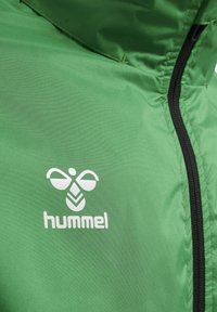 Zielona kurtka z czarnym zamkiem błyskawicznym, z białym logo hummel i emblematem pszczoły na lewej piersi.