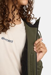 Olijfgroene jack met zwarte accenten, met een ritssluiting en zachte stof. Zichtbaar logo op een lichtbeige t-shirt eronder.