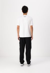 Tricou alb cu mânecă scurtă, guler rotund, cu un logo negru "Barbour" pe spate, purtat cu pantaloni cargo negri și adidași gri.