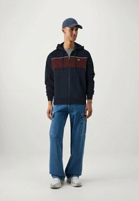 Navy zip-up hoodie with maroon horizontal stripe, paired with light blue wide-leg jeans. Model wears a matching navy cap and white sneakers.