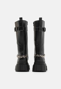 Schwarze kniehohe Stiefel aus Kunstleder mit einer klobigen Plattform, verstellbaren Riemen und einem silbernen Kettenakzent um den Knöchel.