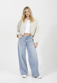 Veste bomber courte de couleur gris clair avec des poignets élastiques, associée à un jean large bleu clair. La mannequin se tient devant un fond blanc.