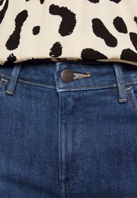 Gros plan de la ceinture d'un jean en denim bleu avec un bouton métallique à côté d’un tissu tacheté noir et blanc glissé à l’intérieur.