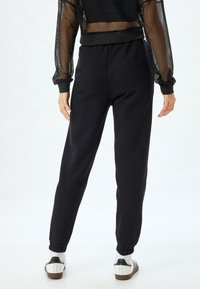 Svarte sweatpants med elastiske manchetter og en afslappet pasform, parret med en sort gennemsigtig mesh langærmet top. Hvide sko med sorte detaljer.