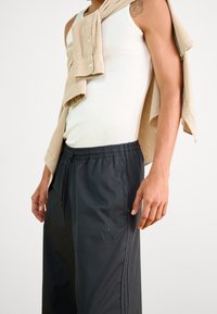 Schwarze Sporthosen mit einem elastischen Bund und seitlichen Taschen, kombiniert mit einem weißen Tanktop und einem beigen Pullover, der über die Schultern gelegt ist.