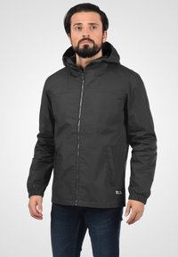 Veste imperméable noire avec une fermeture éclair, des poignets élastiques et une capuche. Elle est fabriquée en tissu lisse, dotée de deux poches latérales et d'un petit étiquette de logo.