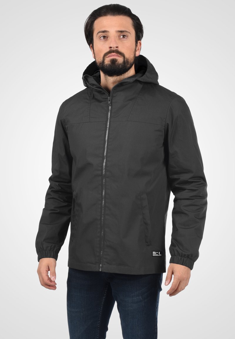 Veste imperméable noire avec une fermeture éclair, des poignets élastiques et une capuche. Elle est fabriquée en tissu lisse, dotée de deux poches latérales et d'un petit étiquette de logo.