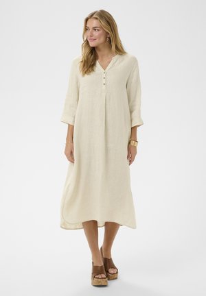 Nő, laza, hosszú bézs ruhában háromnegyedes ujjakkal és barna platform szandálban, egyszínű világos háttér előtt állva.