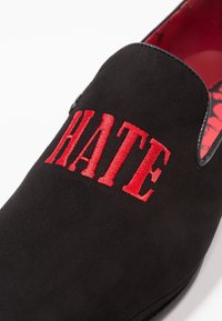 Zwarte suède instapschoen met gedurfde rode geborduurde tekst "HATE" op de bovenkant. De binnenkant heeft een contrasterende rode voering.