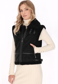 Zwarte nepbont gilet met een rits aan de voorkant, brede kraag en meerdere drukknoopaccenten. Textuurfinish contrasteert met de gladde stof.