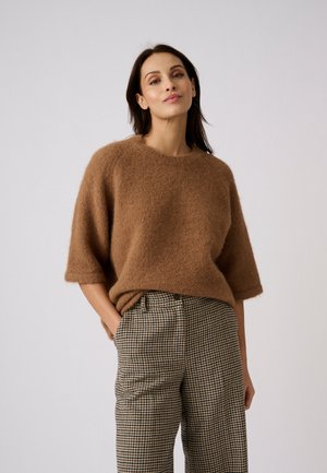 Vrouw die een bruine, pluizige korte mouwen trui en hoog getailleerde geruite broek draagt, staand met één hand in de zak tegen een effen achtergrond.
