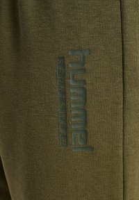 Pantalones deportivos de color verde oliva hechos de tela acanalada, con un gran logo en relieve y texto en el costado como marca.