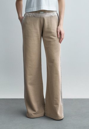 Osoba ubrana w beżowe spodnie z szerokimi nogawkami, z szarymi bocznymi panelami i elastyczną talią oznaczoną napisem "OFF", stojąca na szarej podłodze na tle jednolitej ściany.