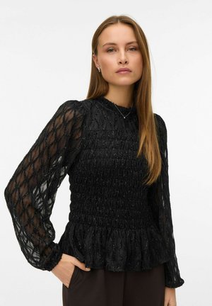Frau mit langen braunen Haaren, die eine schwarze, strukturierte, transparente Bluse mit langen, aufgeblähten Ärmeln und hohem Kragen trägt und mit einer Hand in der Tasche posiert.