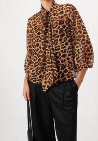 Blouse à motif léopard avec un détail à nouer, fabriquée en tissu transparent. Présente des manches bouffantes et une coupe décontractée, associée à un pantalon noir taille haute.