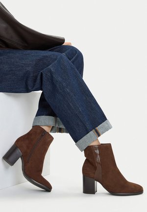 Brązowe botki zamszowe za kostkę na obcasie typu słupek, z bocznym zamkiem i błyszczącymi skórzanymi akcentami. Noszone z podwiniętymi ciemnymi spodniami dżinsowymi.