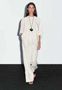 Un mannequin marche avec confiance dans une tenue gris clair composée d'un haut ample et de pantalons larges, accessoirisée d'un collier en pendentif en forme de cœur noir.