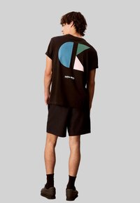 Schwarzes T-Shirt mit einem großen Grafikmotiv in Blau, Türkis, Pink und Weiß. Kombiniert mit schwarzen Shorts und dunklen Schuhen. Einfaches, lässiges Design.