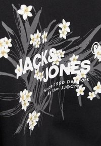 Tejido negro con un diseño floral que presenta flores blancas y amarillas, junto al texto "JACK & JONES" en tipografía gruesa blanca.