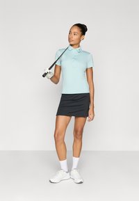 Ljusblå golfpolo med korta ärmar, svart golfficka och vita sneakers; modellen håller i en golfklubba; enkel, strömlinjeformad design.